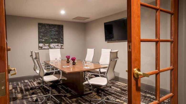 A modern conference room with a wooden table, white chairs, a wall-mounted TV, and artwork. The room has soft lighting and a patterned carpet.