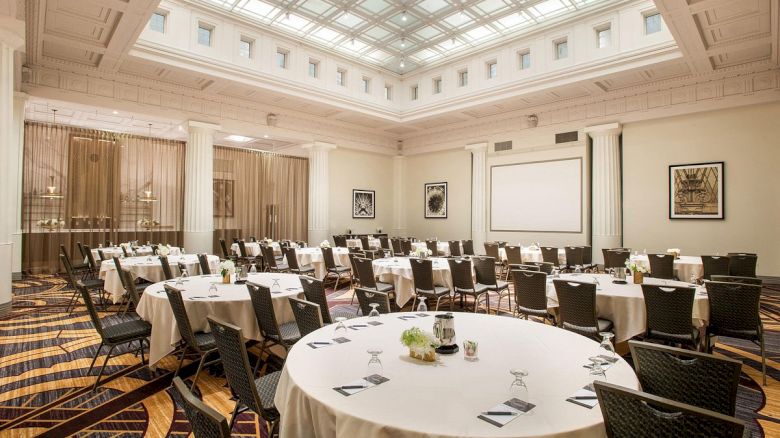 The image shows a conference room with round tables, chairs, a large screen, and high ceiling. The room is set up for a meeting or event.