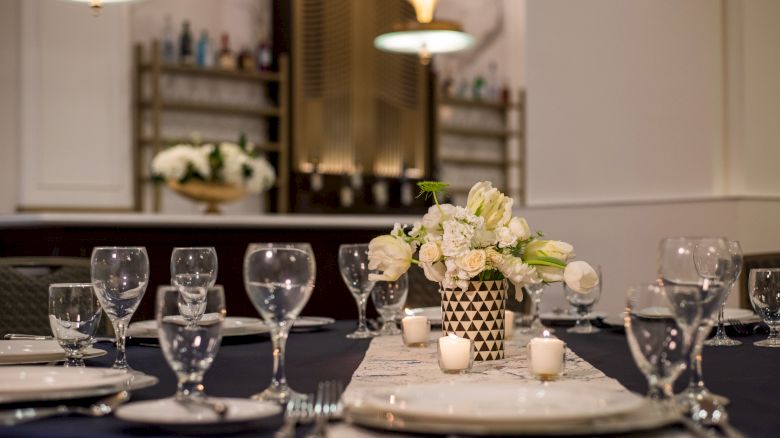 A dining table set elegantly with glassware, plates, and a floral centerpiece, in a room with soft lighting and a decorative background.