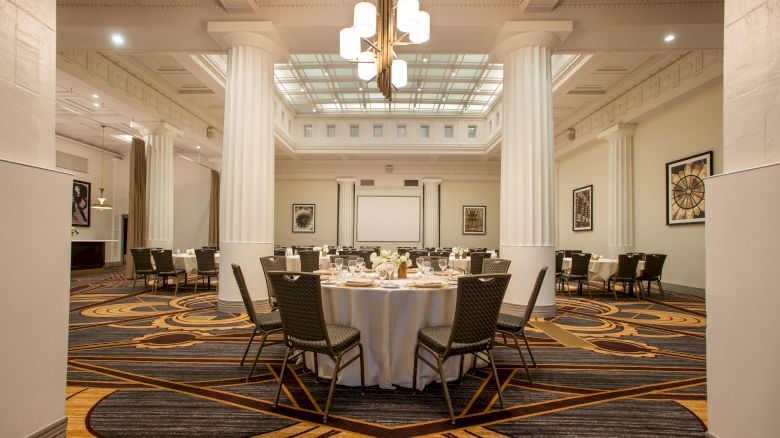 A spacious dining hall with round tables set for a formal event, featuring elegant lighting and decorative columns.