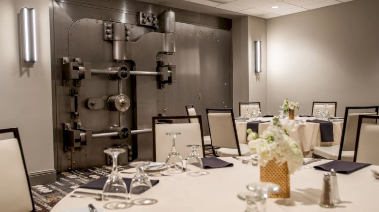 A dining area set against a large bank vault door, featuring elegantly arranged tables with glasses and napkins.