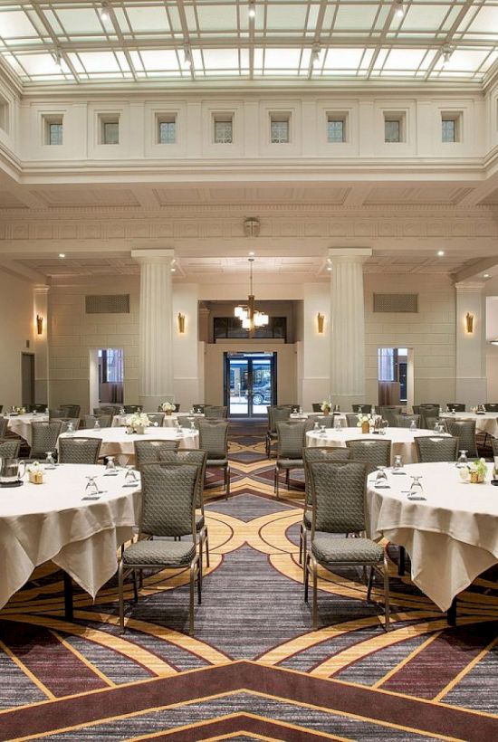 A spacious banquet hall with round tables set for dining, elegant decor, and a large skylight above, creating an inviting atmosphere.