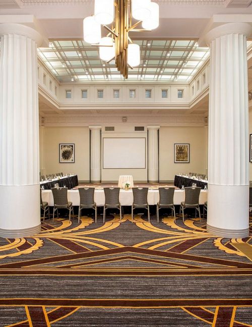 A spacious conference room with elegant columns, a projector screen, and chairs arranged around a table, featuring decorative carpeting.