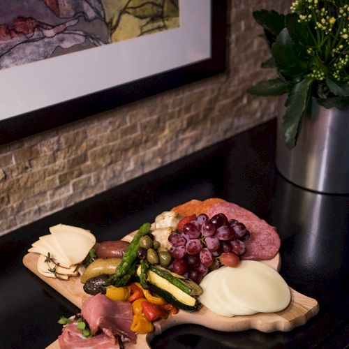 A charcuterie board with assorted meats, cheeses, and grapes on a table next to a potted plant and framed artwork on the wall.