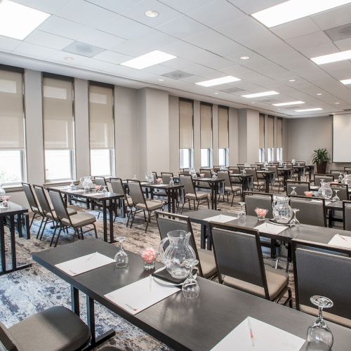 The image shows a conference room setup with rows of tables, chairs, notepads, and glasses, facing a projector screen in a well-lit space.