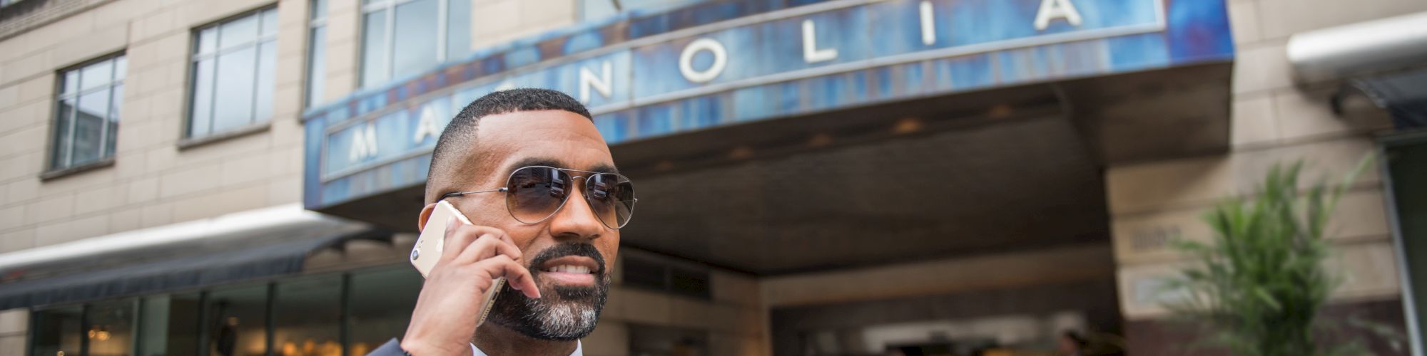 A man in a suit and sunglasses talks on the phone in front of the Magnolia hotel entrance with flags above.