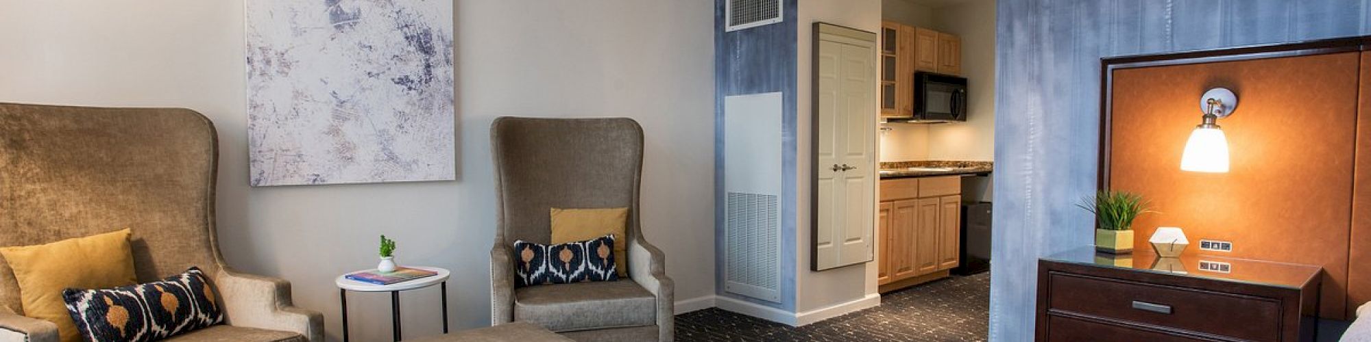 The image shows a hotel room with two armchairs, an ottoman, a dresser, and a kitchenette, featuring modern decor and dark accents.