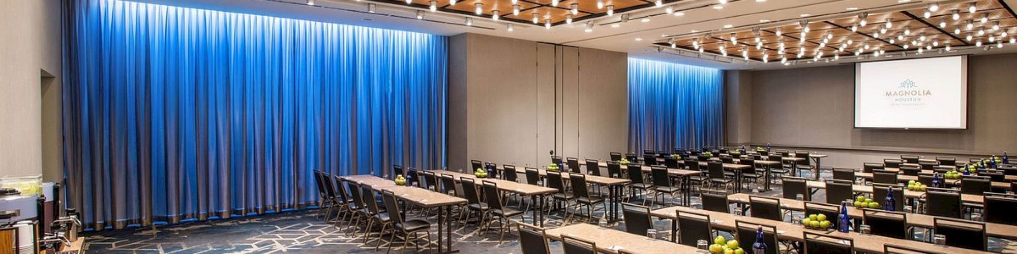 A conference room with rows of tables and chairs, blue curtains, lighting on the ceiling, and a screen displaying 