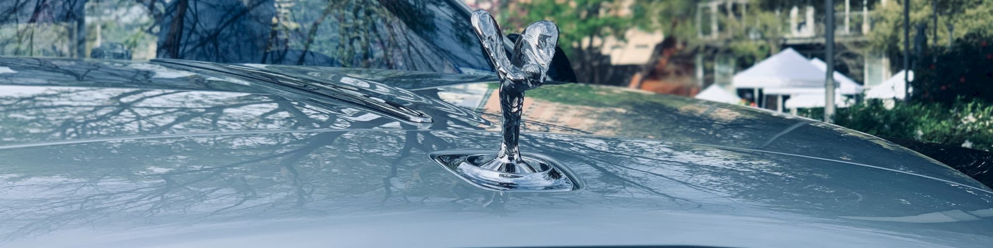 The image shows the hood and emblem of a Rolls-Royce car with the Spirit of Ecstasy ornament in an outdoor setting.