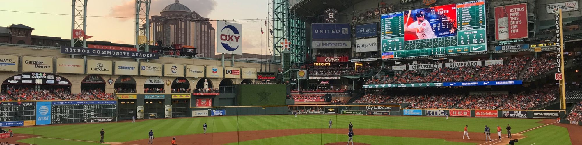 A baseball game in a stadium with a large crowd, featuring a clear sky and a partially retractable roof in the background.