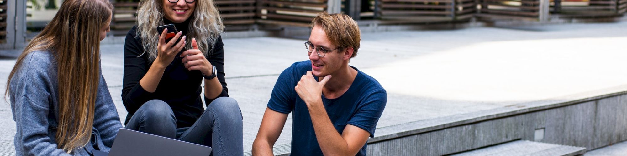 Three people sitting on steps, one with a laptop, two others engaged in conversation and smiling, with a building facade in the background.