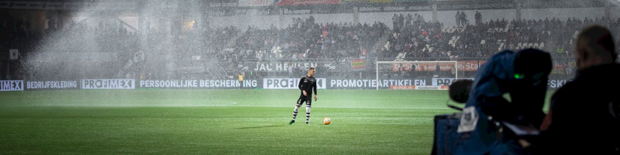 A soccer player stands on a field under stadium sprinklers, with cameras and spectators in the background.