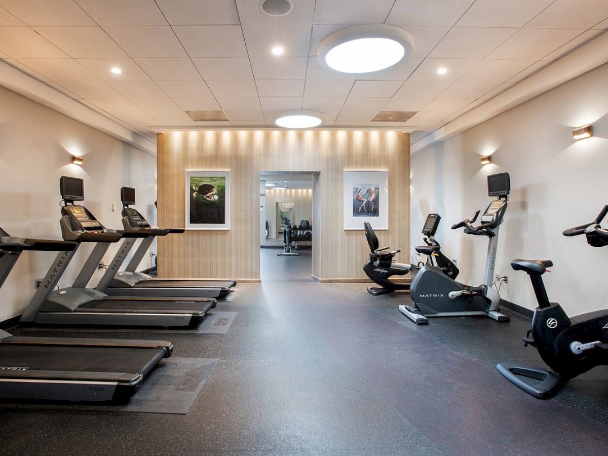 The image shows a gym with treadmills, stationary bikes, and wall-mounted TVs in a well-lit room with framed pictures.