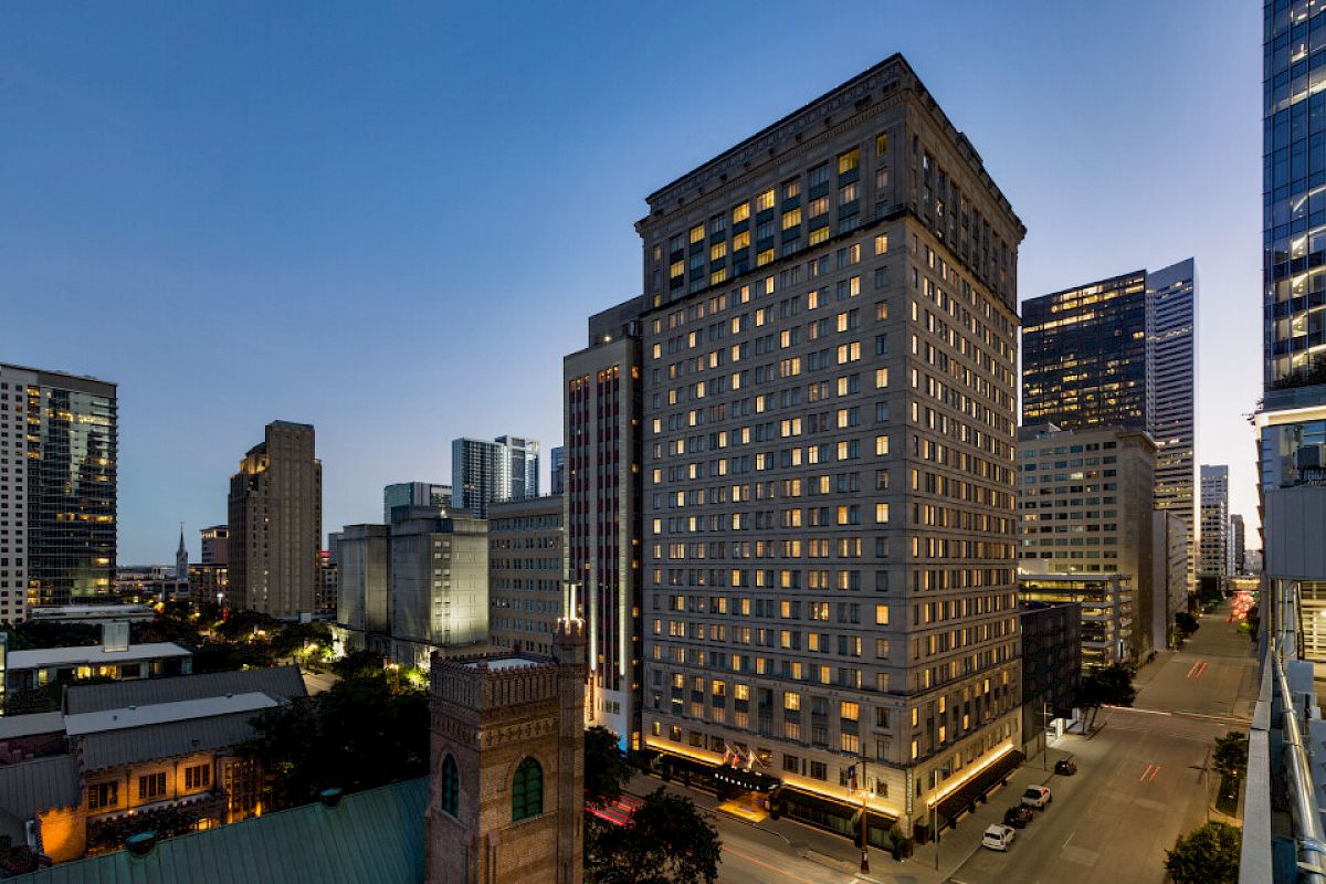 A cityscape at dusk featuring tall buildings, a street view, and a clear sky. The scene includes lit windows and sparse traffic.