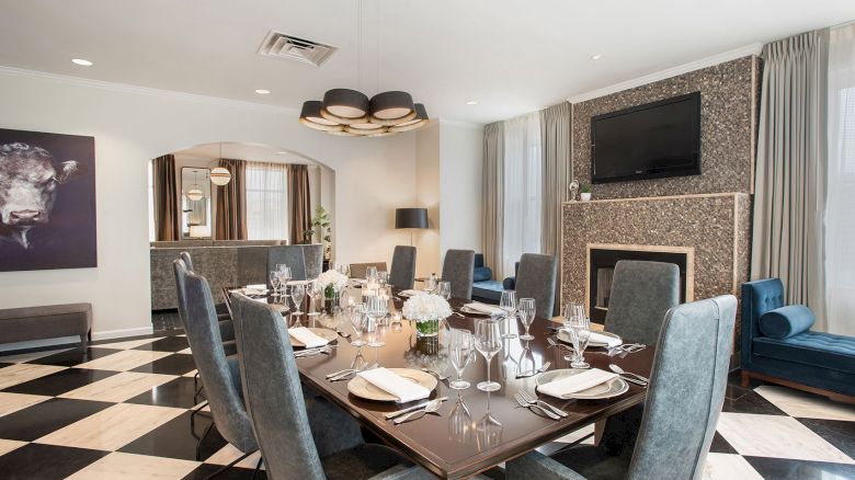 Elegant dining room with a set table, plush chairs, checkered floor, modern lighting, a painting, and a fireplace with a TV above it.