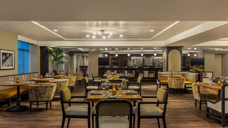 The image shows a modern restaurant interior with neatly arranged tables and chairs, elegant decor, and a bar area in the background.