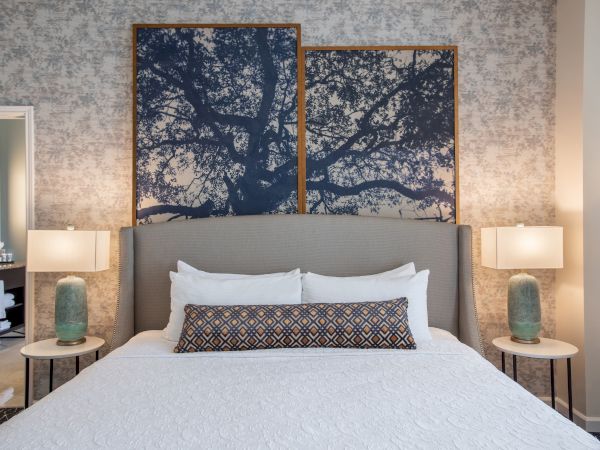 A modern bedroom features a large bed with white linens, flanked by two lamps on side tables, and abstract tree artwork above the headboard.