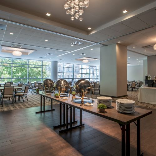 A modern dining area with a buffet table set with chafing dishes, plates, and cutlery under a decorative ceiling light.