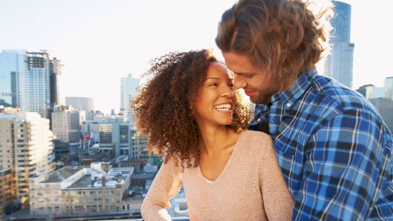 A couple embraces and smiles at each other on a city rooftop, surrounded by tall buildings and a clear sky.