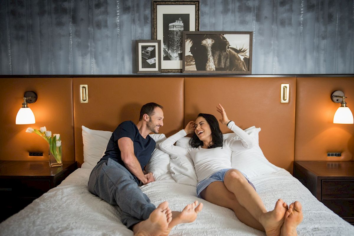 A couple is relaxing on a bed in a warmly lit room, with framed artwork above them and a vase of white flowers on the side table.