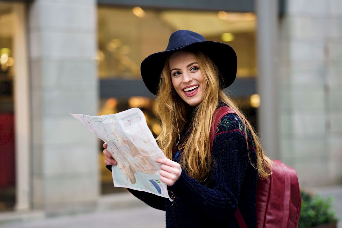 A smiling person with long hair and a hat, holding a map, stands on a street with shops in the background, wearing a backpack.