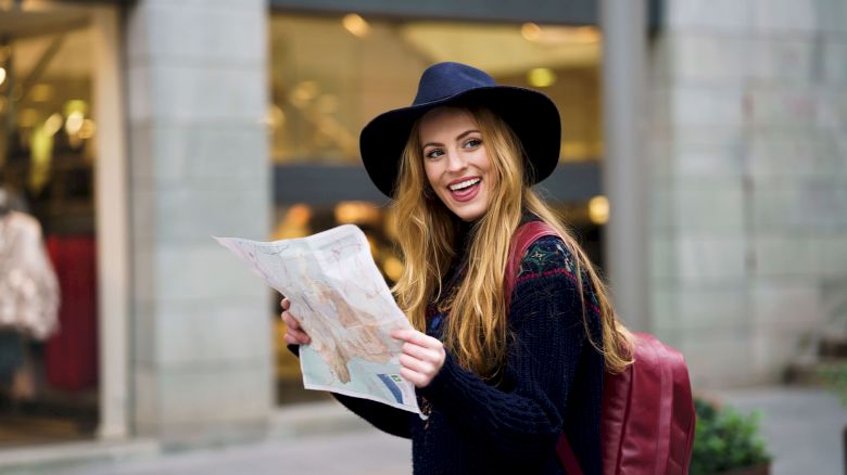 A smiling person with long hair and a hat, holding a map, stands on a street with shops in the background, wearing a backpack.