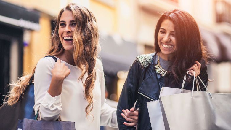 Two women are walking outdoors, smiling, and carrying shopping bags. They're enjoying a sunny day in a city setting.