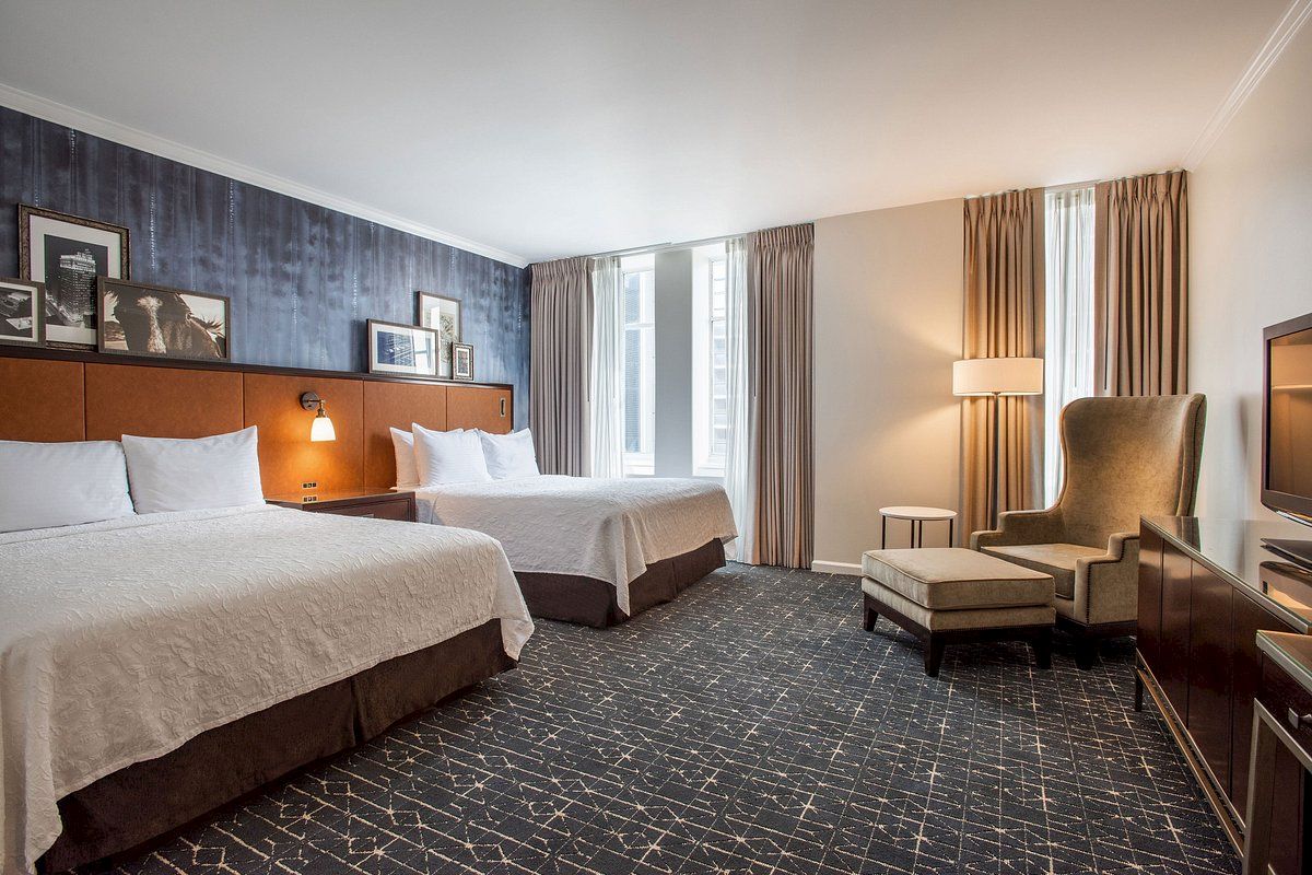 A hotel room with two beds, a cozy chair, a lamp, framed pictures, and curtains, creating a warm and inviting atmosphere.