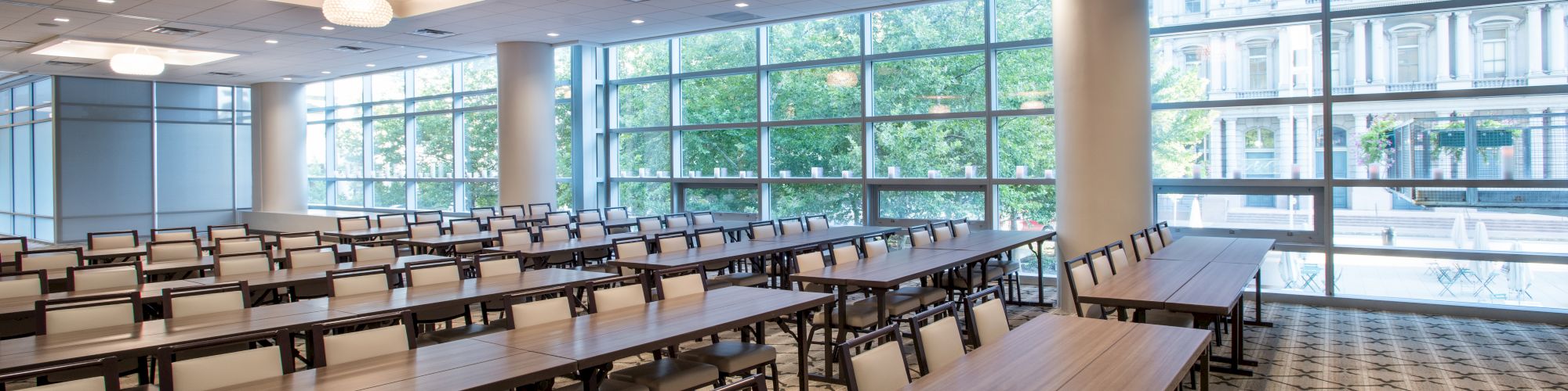 A modern conference room with rows of empty tables and chairs, large windows, and ceiling lights, offering a spacious and bright atmosphere.