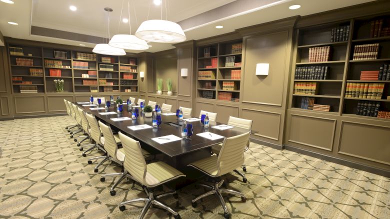 A conference room with a long table, surrounded by chairs, bookshelves, and notepads with water bottles on the table enhances the setting.