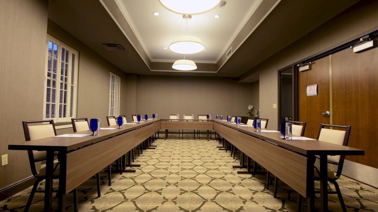 The image shows a conference room with U-shaped tables, chairs, and blue glasses, featuring overhead lighting and patterned carpet.