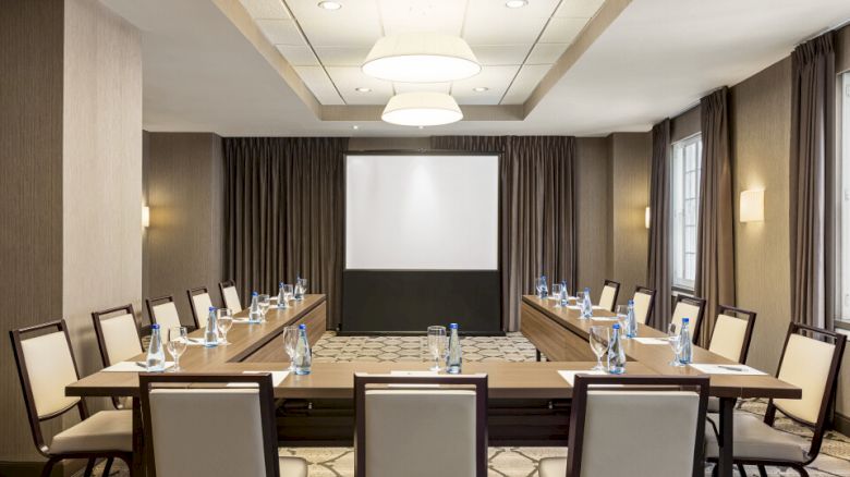 A conference room with a U-shaped table setup, chairs, a projector screen, water bottles, and note pads in a well-lit space.