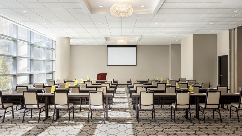 A conference room with rows of chairs, tables, a projector screen, and natural light from large windows, ready for a presentation.