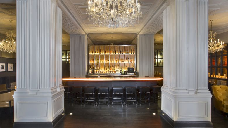 A modern bar with elegant chandeliers, tall white columns, and a dimly lit atmosphere, featuring a well-stocked backlit bar.