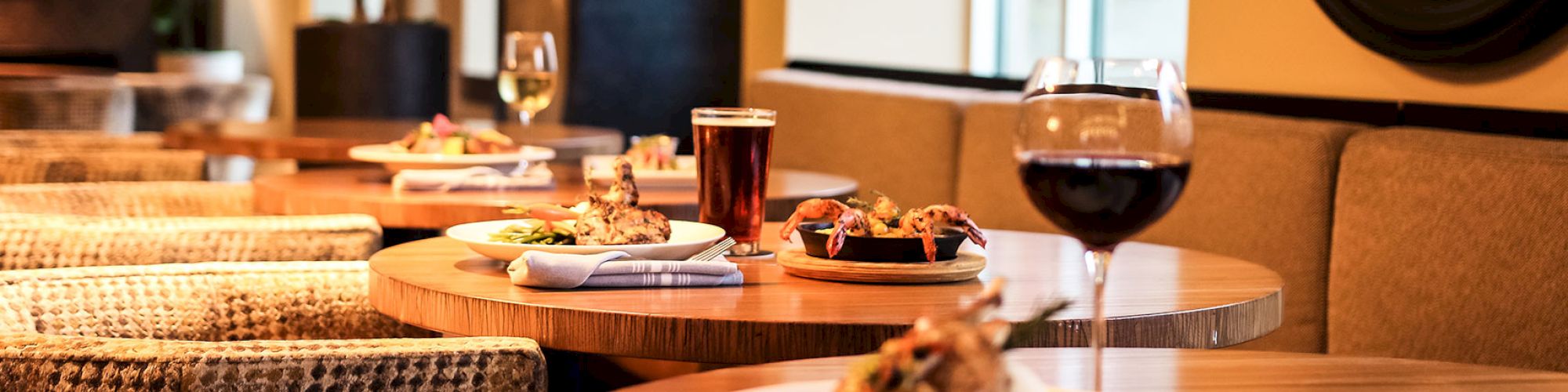 A cozy restaurant scene with tables set with wine, beer, and plates of food in a warmly lit atmosphere.