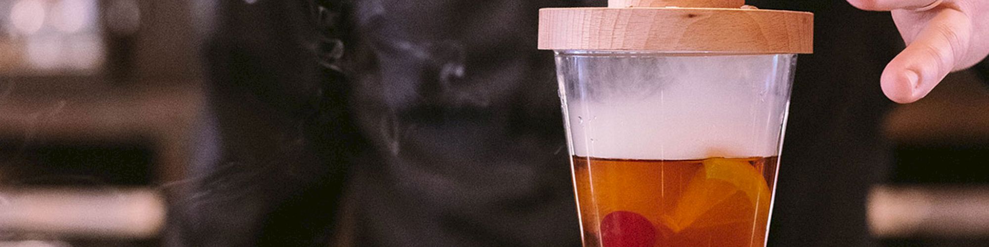 A hand lifts a wooden lid from a smoky cocktail on a bar counter, revealing a drink with a cherry and citrus twist inside.