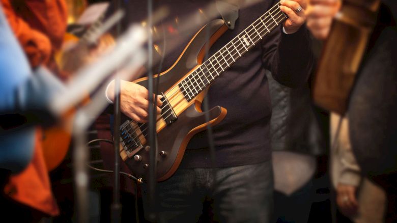 A person is playing an electric bass guitar during a performance. Other musicians and instruments are partially visible around them.