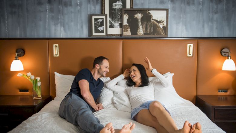 A couple is relaxing on a bed, smiling at each other in a warmly lit room with artwork and lamps on the wall.