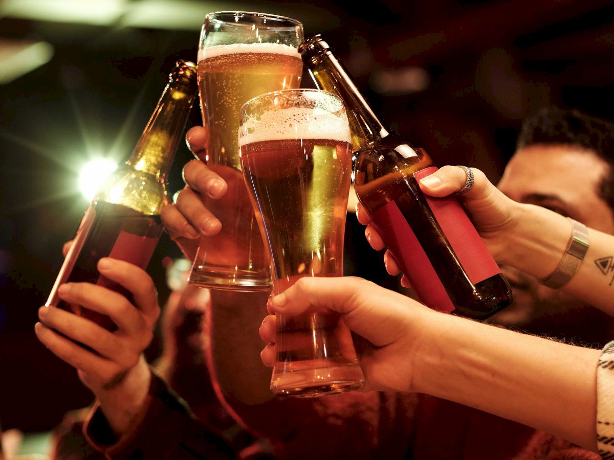 People clinking beer glasses and bottles together at a table in a warm, dimly lit setting, celebrating or making a toast.