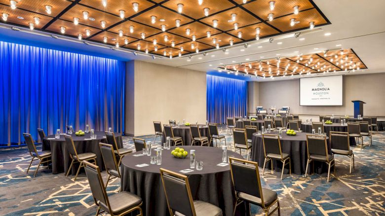 The image shows a modern conference room with round tables, chairs, a podium, overhead lights, and a screen displaying 