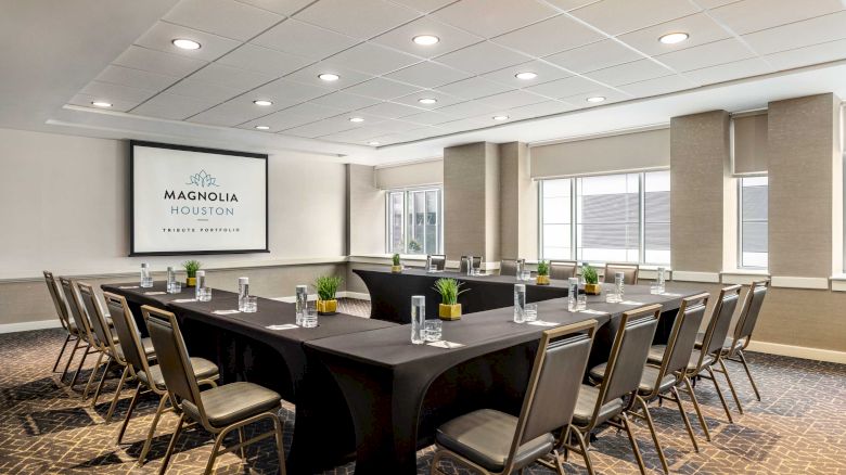 A conference room with a U-shaped table setup, chairs, water glasses, and a screen displaying 