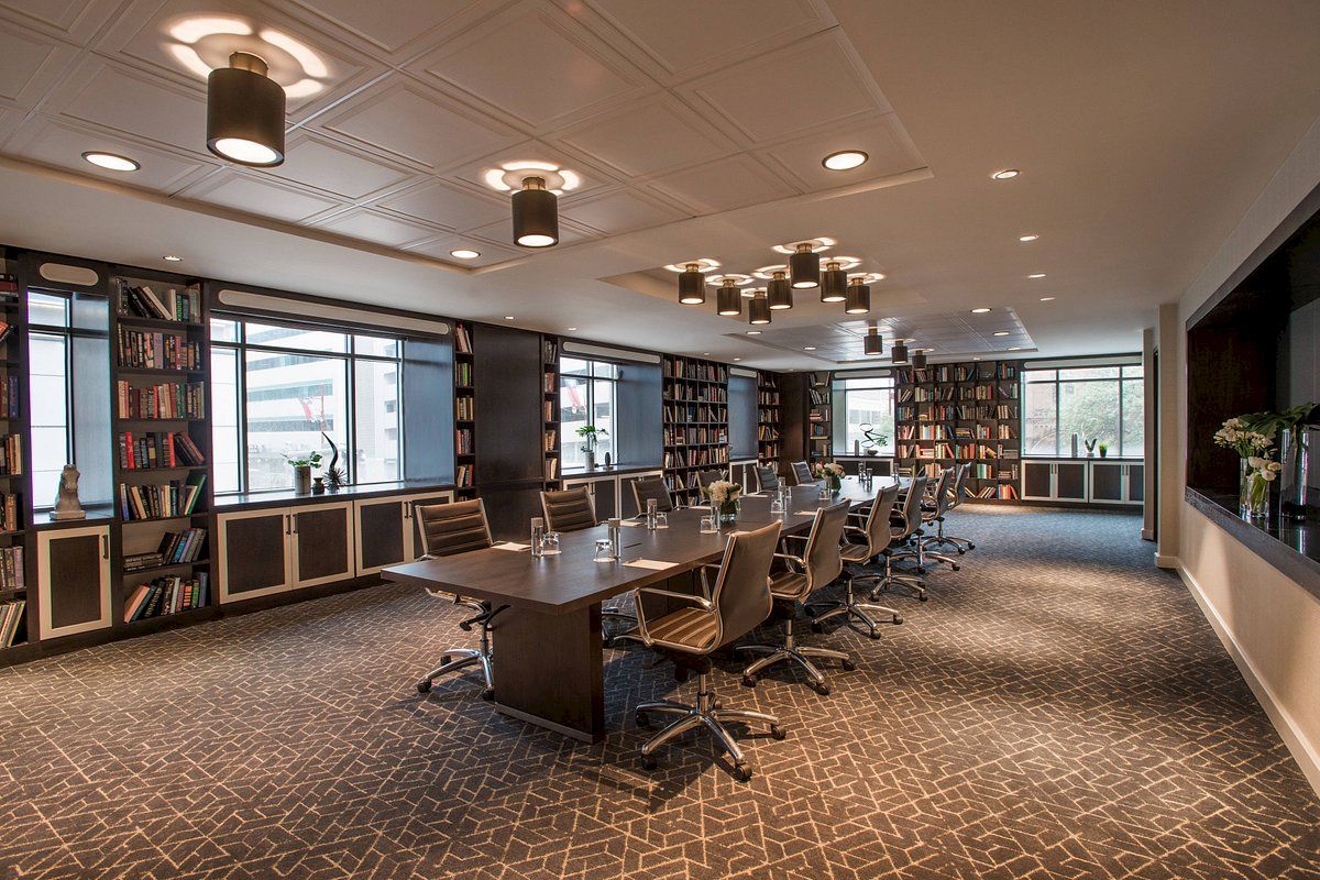 The image shows a modern conference room with a long table, swivel chairs, bookshelves, and large windows letting in natural light.