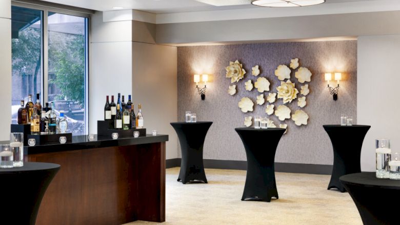 This image shows an elegant room setup for a reception with high-top tables, a bar with wine bottles, and floral wall decorations.