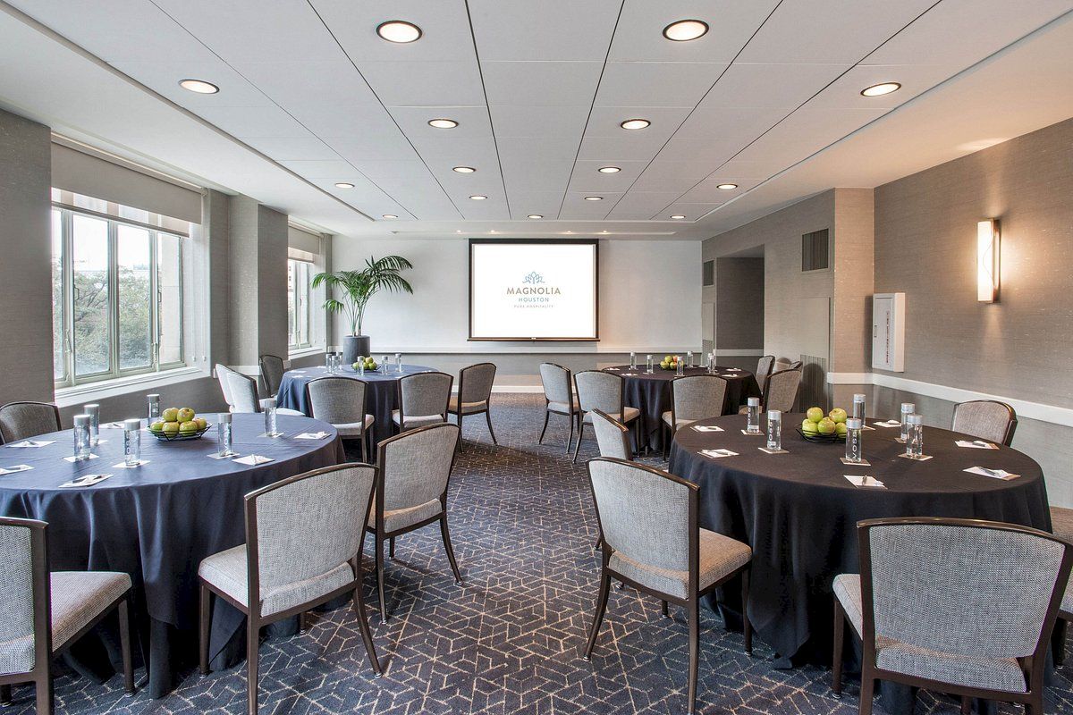 A conference room with round tables, chairs, a projector screen, and windows. Tables have water glasses and apples.