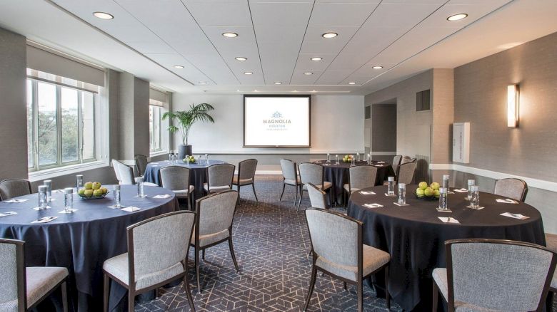 A conference room with round tables, chairs, a projector screen, and windows. Tables have water glasses and apples.