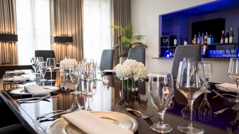 A dining table set elegantly for a formal gathering, with flowers, glassware, and napkins; a blue-lit bar is visible in the background.