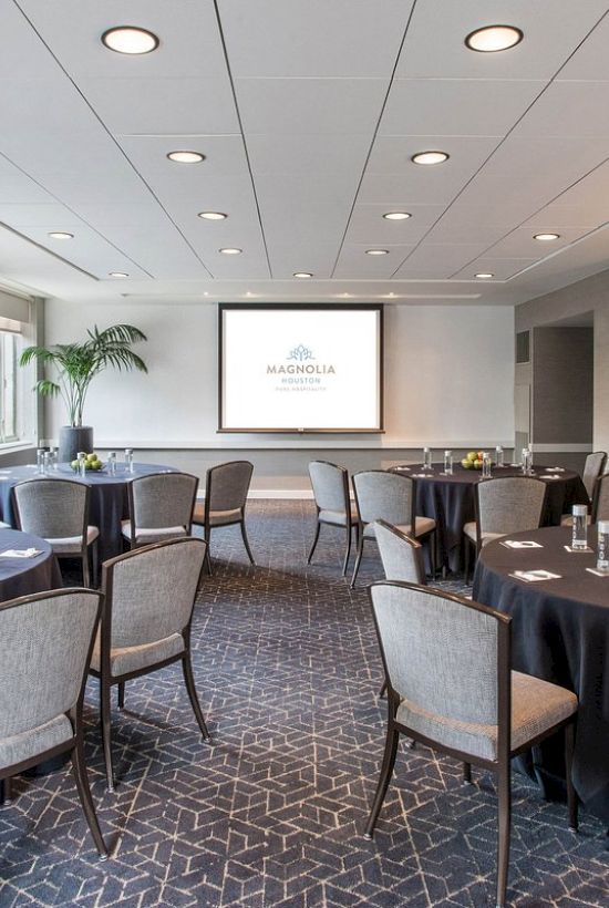 A conference room with round tables, chairs, a projector screen, and natural light from windows.