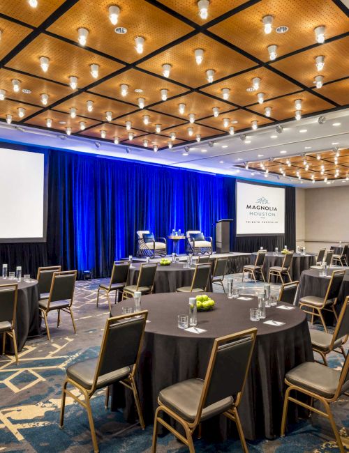 A conference room set up with round tables, chairs, blue curtains, and large screens displaying 