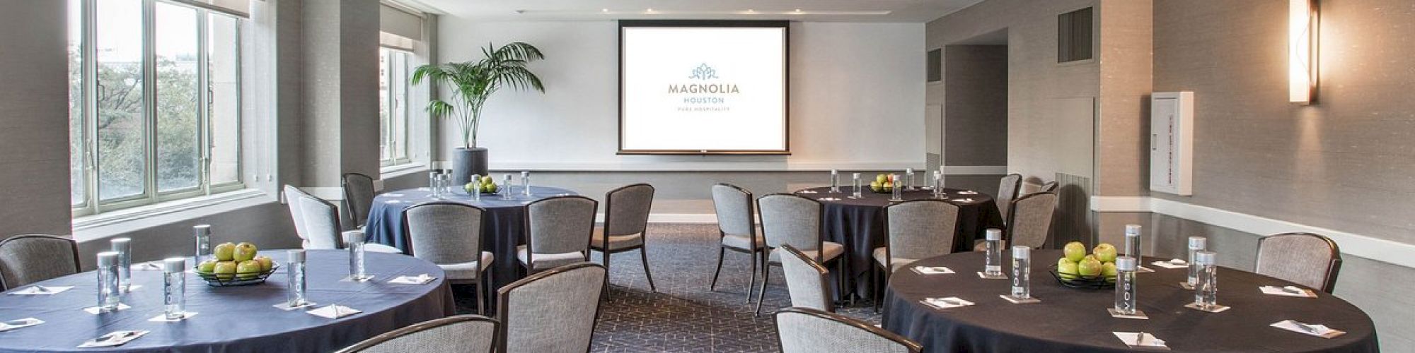 A conference room with round tables, chairs, a projector screen, and natural light from large windows is set up for a meeting or event.