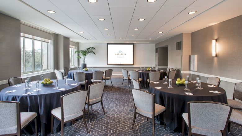 A conference room with round tables, chairs, a screen, and decorative plants. Tables have black cloths and are set for a meeting.